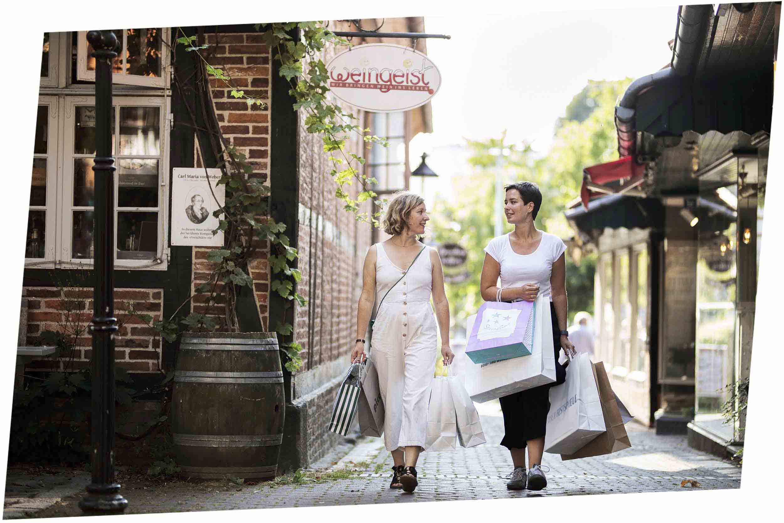 Zwei Frauen mit Einkaufstaschen schlendern durch die historische Altstadt von Eutin, vorbei an Fachwerkhäusern und kleinen Geschäften wie Weingeist.